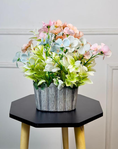 Decorative flower arrangement in a silver pot on a hexagonal black table against a light gray wall.