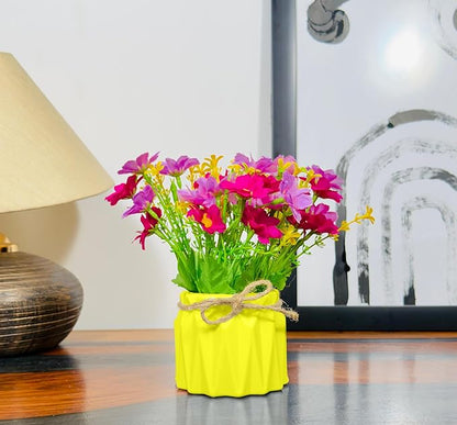 Colorful artificial flowers in a yellow vase on a wooden surface with a lamp and abstract painting in the background.
