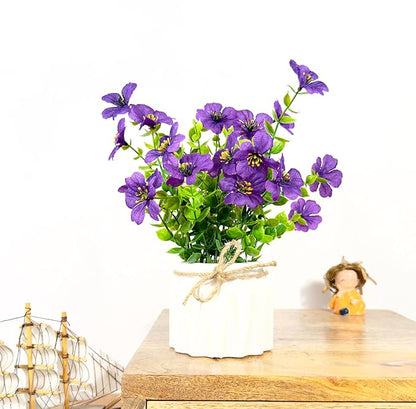 Purple artificial flowers in a white pot on a wooden surface with a small figurine and decorative ship in the background.