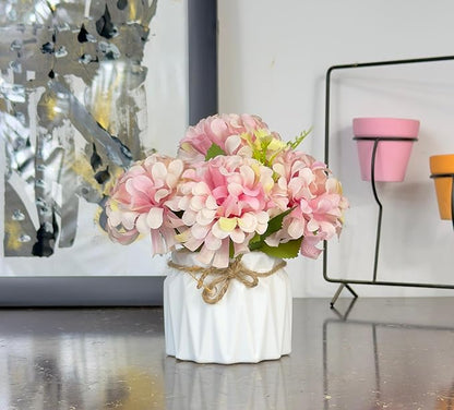 Pink floral arrangement in a white vase on a reflective surface with decorative elements in the background.