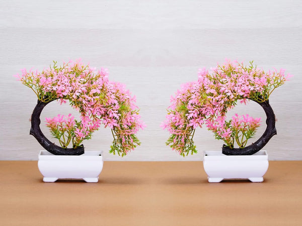 Two artificial plastic faux plants with pink flowers, displayed in white pots, with a black circular base supporting each plant.